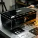 Elegante schwarze Automotive-Geschenkbox mit Chromakzenten auf polierter Stahlwerkbank, umgeben von Druckmustern mit geprägten Logos