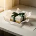 Elegante Geschenkbox mit grünem Band, Seidenpapier und handgeschriebener Karte auf weißem Tisch im warmen Licht.