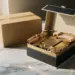 Offene magnetische Geschenkbox mit Seidenpapier und Premium-Produkt neben braunem Versandkarton auf Marmoroberfläche.