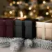 Drei elegante Geschenkboxen in Burgunderrot, Mattschwarz und Champagnergold auf Tannenzweigen mit warmem Kerzenlicht im Hintergrund.