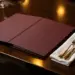 Aufgeschlagene kratzfeste Speisekarte auf poliertem dunklem Holztisch mit Leinenserviette und elegantem Besteck im Kerzenlicht.