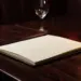 Aufgeschlagene Ledermenümappe auf poliertem Walnusstisch im Restaurant, cremefarbene Seiten im warmen Kerzenlicht, Weinglas unscharf im Hintergrund.