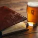 Ledergebundene Speisekarte mit Hopfen-Prägung auf rustikalem Eichentisch neben frisch gezapftem Amber Bier im Kerzenlicht.