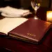 Aufgeschlagene Ledermappe auf poliertem Holztisch im Restaurant, Kerzenlicht beleuchtet den geprägten Einband, Weinglas im Hintergrund.