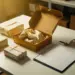 Luxuriöse Geschenkbox halb geöffnet mit Seidenpapier, umgeben von verpackten Produkten und Versand-Checkliste auf weißem Lagertisch.