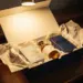 Geöffnete luxuriöse Geschenkbox mit Premium-Produktproben auf poliertem Eichenschreibtisch, beleuchtet von warmem Spotlight in Creme- und Marineblautönen.
