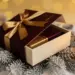 Luxuriöse Geschenkbox mit Satinband, halb geöffnet auf Tannenzweigen, warmes Kerzenlicht, Burgunderrot und Gold.