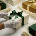 Behandschuhte Hände schließen eine luxuriöse Geschenkbox mit Satinband, umgeben von edlem Verpackungsmaterial in Waldgrün, Gold und Elfenbein.