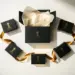 Offene matte Geschenkbox in Schwarz-Gold mit fünf eleganten Produktverpackungen im Halbkreis, Seidenpapier und goldenen Bändern auf weißem Untergrund.