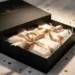 Luxuriöse matte Geschenkbox mit halbgeöffnetem Deckel, Produkte in Seidenpapier und Satinband auf weißem Marmor mit Goldkonfetti.