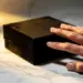 Schwarze Luxus-Geschenkbox mit samtiger Oberfläche wird von Fingerspitzen berührt auf weißem Marmor mit goldener Beleuchtung