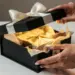 Elegante Hände öffnen eine matte schwarze Geschenkbox mit weißem Band, aus der warmes goldenes Licht und Seidenpapier hervorscheinen.
