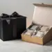Zwei elegante Geschenkboxen auf Marmor – eine mattschwarze Box mit Satinband, eine geöffnete Box mit Produktproben in Seidenpapier.