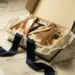 Geöffnete Luxus-PR-Box auf Marmor mit Seidenkleidung, Parfümflakon, Seidenpapier und marineblauem Band im goldenen Studiolicht.