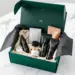 Geöffnete smaragdgrüne PR-Box mit Premium-Lifestyle-Produkten auf weißem Marmortisch, Studio-Beleuchtung