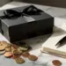 Matte schwarze Geschenkbox mit Satinband auf Marmor, umgeben von Euromünzen und aufgeschlagenem Notizbuch im warmen Licht.