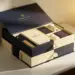 Gestapelte Onboarding-Boxen auf einem Schreibtisch, die oberste geöffnet mit Notizbüchern und Willkommensartikeln, in Navy und Cremeweiß mit Goldakzenten.