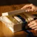 Elegante Hände öffnen eine Premium-Geschenkbox mit Seidenpapier und Satinband in Creme und Dunkelblau auf Eichenholzoberfläche.