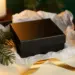 Matte schwarze Geschenkbox mit geprägtem Logo auf weißem Seidenpapier, dekoriert mit Tannenzweig und goldener Schleife.