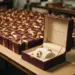 Produktionstisch mit gestapelten burgunderroten Premium-Geschenkboxen, goldenen Schleifen und geöffneter Box mit luxuriösem Innenleben im Vordergrund.