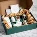 Geöffnete grüne Premium-PR-Box mit goldenen Akzenten zeigt Kaffee, Wasserflasche, Kopfhörer und Snacks auf Marmortisch