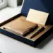 Aufgeklappte Onboarding-Box mit Leder-Notizbuch, Stift und Willkommenskarte auf weißem Schreibtisch in Navy und Champagnergold.