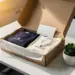 Geöffnete Onboarding-Box mit Firmenmerchandise, Willkommentskarte und Sukkulente auf weißem Schreibtisch in warmem Licht.