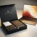 Schwarze Onboarding-Box mit geöffnetem Deckel, Willkommensmaterialien, USB-Karte und Notizbuch, neben Laptop mit leuchtendem Willkommensbildschirm.