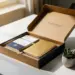 Geöffnete Onboarding-Box mit Notizbuch, Willkommensmaterialien und kleiner Sukkulente auf weißem Schreibtisch im warmen Licht.