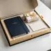 Geöffnete Premium-Onboarding-Box mit Notizbuch, Willkommenskarte und Geschenk in Elfenbein, Marineblau und Gold auf weißem Schreibtisch.