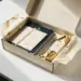 Geöffnete Onboarding-Box mit Notizbuch, Stift, Grußkarte und verpackten Merchandise-Artikeln in Creme, Marineblau und Gold auf weißem Schreibtisch.