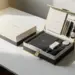 Zwei Premium-Onboarding-Boxen auf weißem Schreibtisch: eine kompakt, eine groß mit Notizbüchern und Accessoires in Elfenbein, Anthrazit und Gold.