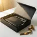 Offene matte Onboarding-Geschenkbox in Schwarz mit monogrammiertem Notizbuch, Stift und goldener Schleife im warmen Abendlicht.