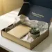 Geöffnete Premium-Onboarding-Willkommensbox mit Notizbuch, Seidenpapier und Sukkulente in Champagner- und Marineblautönen auf weißem Schreibtisch.
