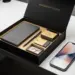 Geöffnete matte schwarze Onboarding-Willkommensbox mit gebrandeten Artikeln auf weißem Schreibtisch, daneben ein Smartphone mit leuchtendem Display.