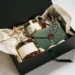 Geöffnete matte schwarze Geschenkbox mit Gourmet-Produkten, Seidenpapier, getrockneten Kräutern und wachsversiegeltem Umschlag auf weißer Fläche.