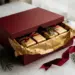 Luxuriöse personalisierte Weihnachtsgeschenkbox mit mattem dunkelrotem Deckel, goldenes Seidenpapier und Tannenzweige sichtbar.