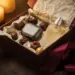Geöffnete Weihnachtsgeschenkbox mit Gourmet-Schokolade, kabellosen Ohrhörern und Parfümflasche in elfenbeinfarbenem Seidenpapier, burgunderfarbenes Band.