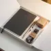 Aufgeklappte Onboarding-Box mit Notizbuch, Stift, Lanyard und Willkommensgeschenk auf weißem Schreibtisch im warmen Licht.