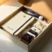 Offene Onboarding-Box mit Notizbuch, Stift, Lanyard und verpackten Willkommensartikeln auf weißem Schreibtisch im warmen Morgenlicht.